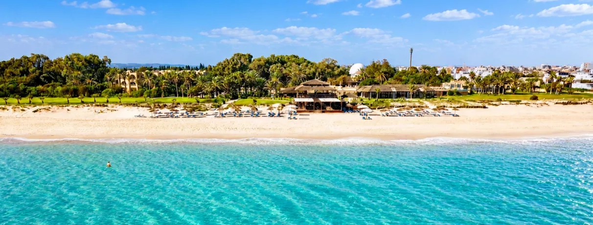 lage de sable avec eau turquoise, transats et palmiers devant un resort entouré de verdure. »