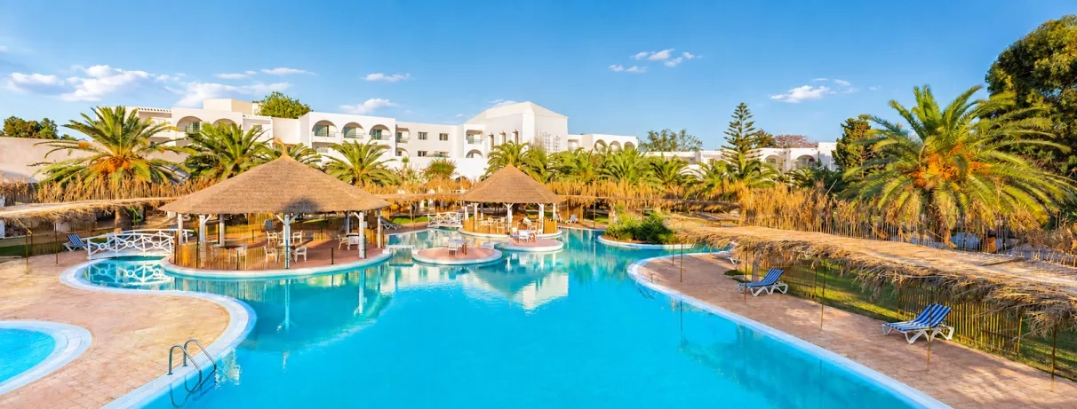 Grande piscine lagon du Mondi Club Shalimar Hammamet bordée de palmiers et d'aillottes en paille.