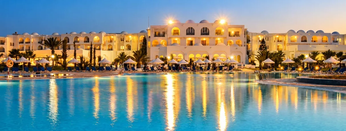 Vue nocturne illuminée de la piscine et des façades blanches de l'hôtel en bord de mer.