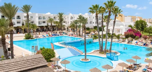 Vue panoramique de la grande piscine extérieure et des bâtiments blancs du Mondi Club Aladin à Djerba, bordée de palmiers et de transats.