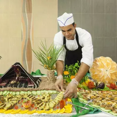 Chef en uniforme préparant un buffet décoratif avec fruits sculptés, légumes et présentation gastronomique.