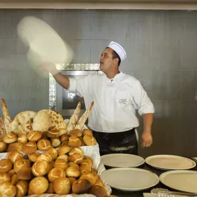 Boulanger en uniforme lançant une pâte à pizza devant un étal de pains variés.