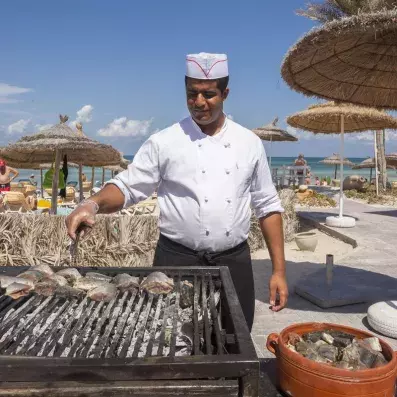 Chef grillant du poisson sur barbecue en plein air près de la plage sous parasols en paille.