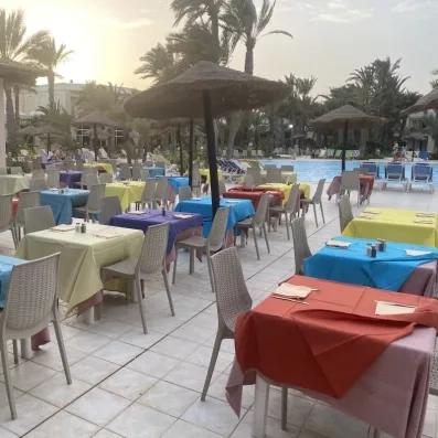 Terrasse du restaurant au bord de la piscine avec tables colorées et parasols en paille, idéale pour un dîner en plein air au coucher du soleil.
