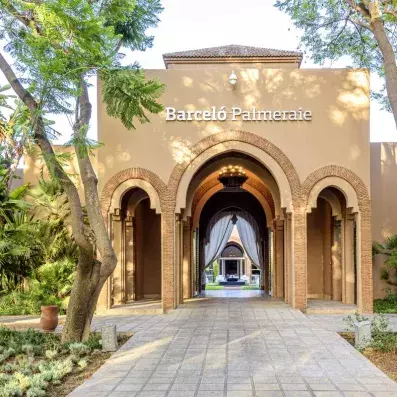L'entrée hôtel avec arches et porte, au fond rideaux et fontaine, en haut lampe et toit, à côté arbres et plantes le long de l’allée