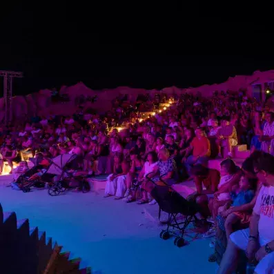 Amphithéâtre extérieur rempli de spectateurs sous éclairage violet pour un spectacle nocturne.