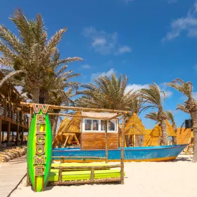 Décor de plage tropicale avec surf ‘Welcome’, cabanes en bois, palmiers et bateau coloré transformé en kiosque, sous un ciel ensoleillé.