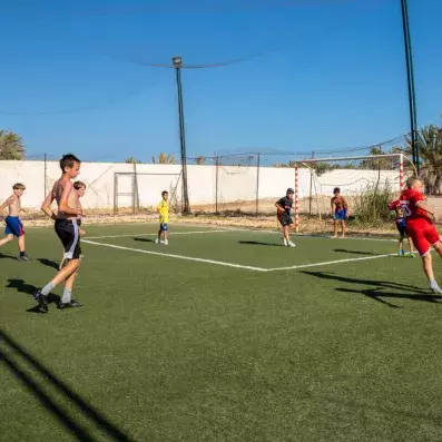 Enfants jouant au football sur terrain synthétique extérieur sous filet de protection.