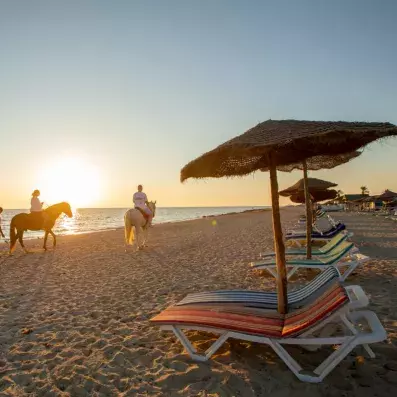 Coucher de soleil sur une plage avec deux personnes à cheval longeant le rivage, transats colorés et parasols en paille au premier plan