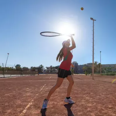 Joueur de tennis frappant la balle sur court extérieur ensoleillé avec palmiers.