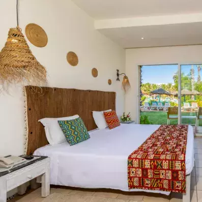 Chambre d’hôtel avec grand lit, décorations murales tressées et vue sur jardin tropical avec transats et palmiers.