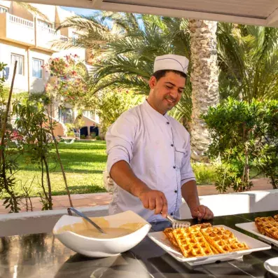 Chef en uniforme préparant des gaufres fraîches dans un jardin tropical ensoleillé.