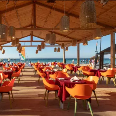 Restaurant en bord de mer avec chaises orange, tables dressées et vue sur la plage.