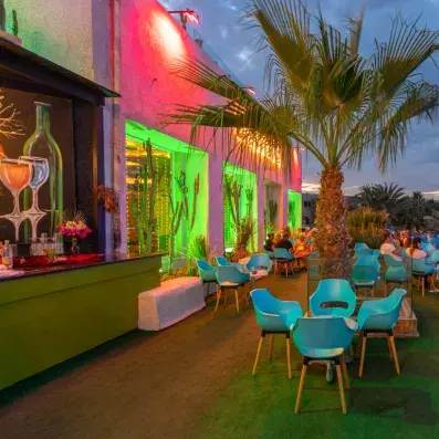 Restaurant nocturne coloré avec éclairages verts et roses, terrasse extérieure et ambiance tropicale.