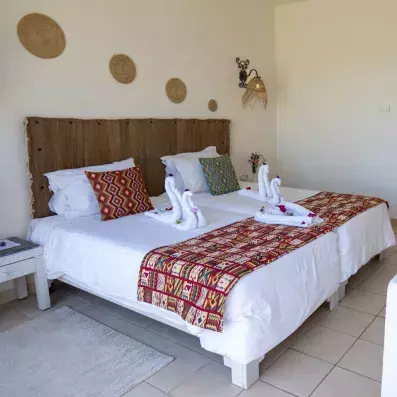 Chambre avec deux lits jumeaux décorés de serviettes en cygnes, coussins colorés et décorations murales en fibres naturelles.