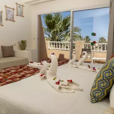 Chambre d’hôtel lumineuse avec lit décoré de serviettes en forme de cygnes, pétales de fleurs et vue sur palmiers depuis le balcon.