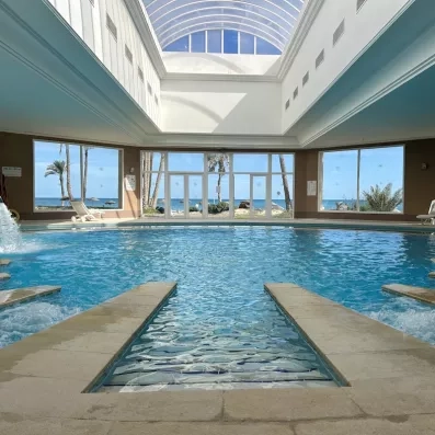Piscine intérieure chauffée sous une coupole en verre lumineuse avec jets massants et vue panoramique sur les jardins et la mer à travers de grandes baies vitrées.
