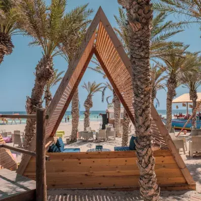 Cabane en bois en A-frame sur plage tropicale avec palmiers, chaises blanches et mer turquoise.