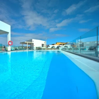 Une vue en perspective de la piscine bordée d'une paroi en verre, sous en ciel bleu.