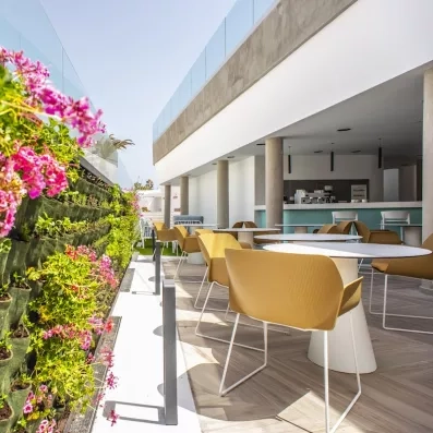 Une terrasse de cafétéria, bordée d'un mur végétal fleuri et meublée de tables rondes.