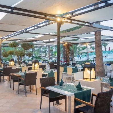 Un restaurant à ciel ouvert sous une pergola ombragée, entre palmiers et piscine, pour dîner dans une ambiance chaleureuse en fin de journée.