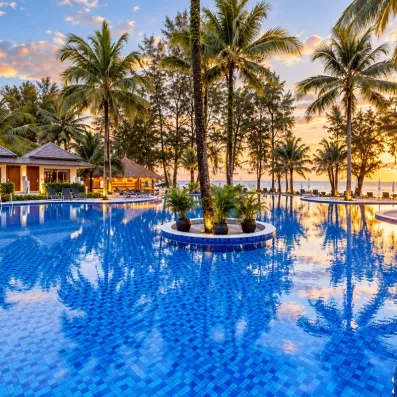 Une large piscine entourée de palmiers et de jardins tropicaux au coucher du soleil.