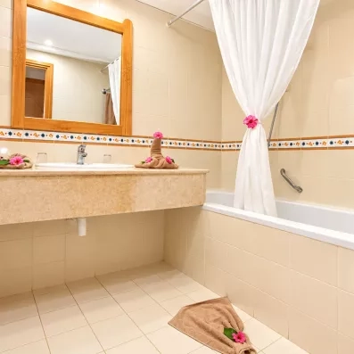 Salle de bain de l'hôtel avec baignoire, grand miroir encadré de bois et décoration soignée avec des fleurs fraîches et des serviettes pliées pour un accueil de bienvenue.