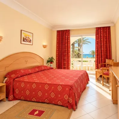 Chambre double lumineuse avec balcon privé offrant une vue sur les palmiers et la mer, équipée d'un mobilier en bois clair et de rideaux rouges chaleureux.