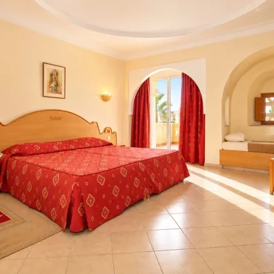 Chambre d'hôtel spacieuse à Zarzis avec un grand lit double couvert d'un couvre-lit rouge typique, une alcôve avec un lit d'appoint et une décoration aux influences tunisiennes.