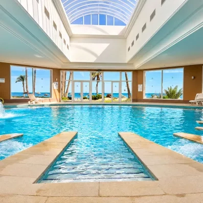 Piscine intérieure chauffée sous une coupole en verre lumineuse avec jets massants et vue panoramique sur les jardins et la mer à travers de grandes baies vitrées.