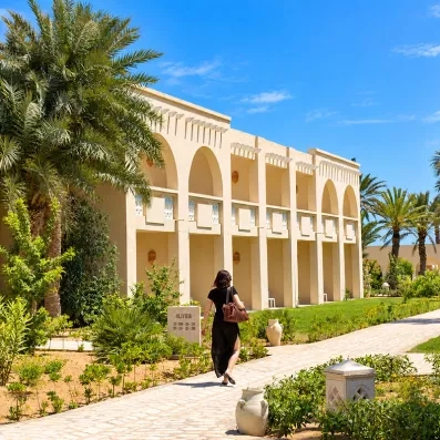 Façade de style architecture tunisienne traditionnelle avec arcades blanches, balcons fleuris et allée piétonne dans les jardins du complexe hôtelier.