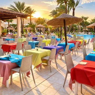 Terrasse du restaurant au bord de la piscine avec tables colorées et parasols en paille, idéale pour un dîner en plein air au coucher du soleil.
