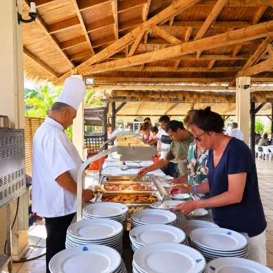 Chef cuisinier servant les clients au buffet de l'hôtel sous une pergola en bois, proposant une variété de plats chauds et spécialités locales.