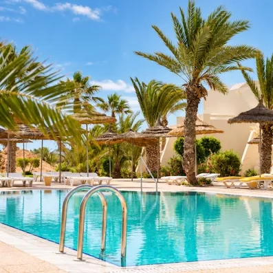 Espace détente avec piscine, parasols en paille et palmiers dans les jardins de l'hôtel à Djerba.