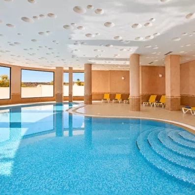 Piscine intérieure chauffée avec transats jaunes et grandes baies vitrées lumineuses à l'hôtel Safira Palms.