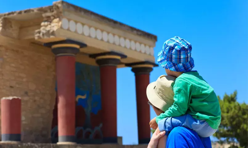 Parent et enfant visitant le palais de Knossos en Crète, colonnes antiques et fresques minoennes sous un ciel ensoleillé.