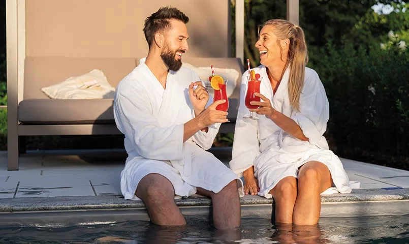 Couple les pieds dans l'eau d'une piscine, en peignoir avec un cocktail dans les mains