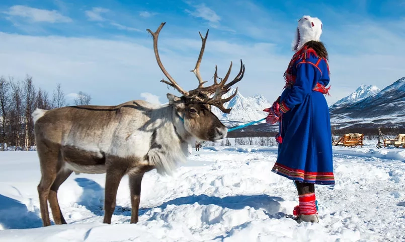 Une personne en costume traditionnel se tient à côté d'un renne dans un paysage enneigé. 