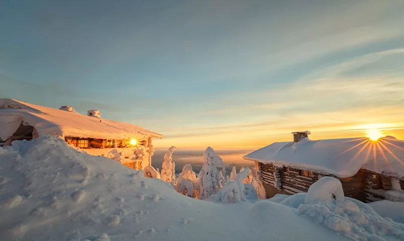 Un lever de soleil sur un paysage enneigé avec des cabanes en bois et des arbres recouverts de neige.