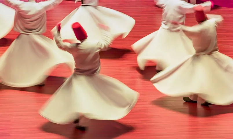 Des derviches tourneurs en robe blanche et chapeau rouge tournent sur une scène rouge. 