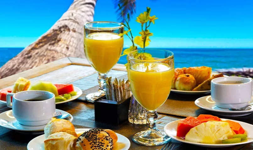 Petit déjeuner sur une table en bois face à la mer, avec des fruits, des viennoiseries et des jus. 